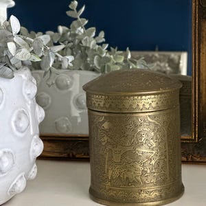 May include: A round, brass container with an intricate, engraved design. The container has a lid and is decorated with a floral pattern and a stylized animal figure.