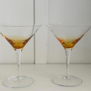 Puede incluir: Dos copas de martini transparentes con cuencos de color ámbar y tallos largos y delgados. Las copas están sobre un fondo blanco, lo que sugiere un bar o un comedor. El diseño es simple y elegante.