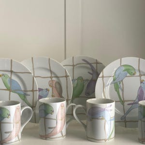 Könnte beinhalten: Ein Set aus vier weißen Keramiktellern und vier passenden Tassen, jeweils verziert mit einem braunen Gittermuster und bunten Illustrationen von Sittichen in Rosa-, Blau-, Grün- und Violetttönen, angeordnet in zwei Reihen zu je vier Objekten.