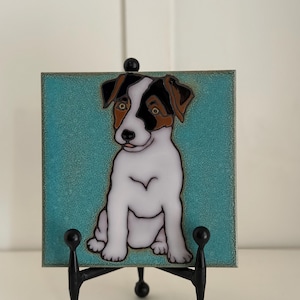 May include: A ceramic tile with a painted image of a Jack Russell Terrier dog on a teal background. The tile is displayed on a black metal easel.