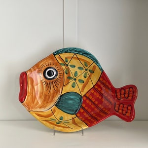 Puede incluir: Un plato de servir de cerámica con forma de pez colorido con un diseño amarillo, naranja, rojo, azul y verde. El pez tiene un ojo grande y un patrón detallado.