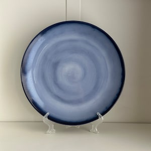 May include: A large, round, blue and white ceramic dinner plate with a swirling pattern. The plate is on a white stand.