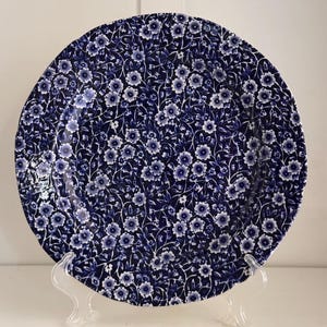 May include: A round, blue and white floral patterned plate. The plate features a dense pattern of small white flowers and leaves against a dark blue background. The plate is displayed on a clear stand.