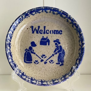 Puede incluir: Un plato de cerámica azul y blanco con un diseño azul y blanco de dos figuras plantando flores frente a una casa. El plato tiene un esmalte azul y blanco moteado y dice "Bienvenido" en letras azules.