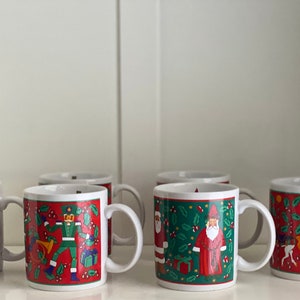 May include: Set of six white ceramic mugs with Christmas-themed designs. Each mug features a different festive scene, including a reindeer, a nutcracker, Santa Claus, and a carousel horse. The mugs have red, green, and yellow accents.