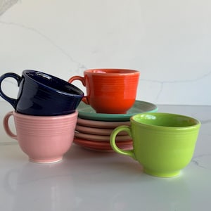 May include: A collection of ceramic cups and saucers in various colors including dark blue, bright orange, light pink, and lime green. The cups feature a rounded shape with a single handle and horizontal ringed detailing around the exterior surface.