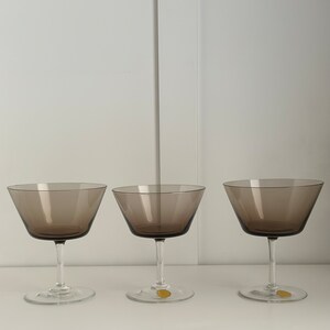 May include: Three clear glass champagne coupes with a dark brown tint. The glasses are empty and sitting on a white surface.