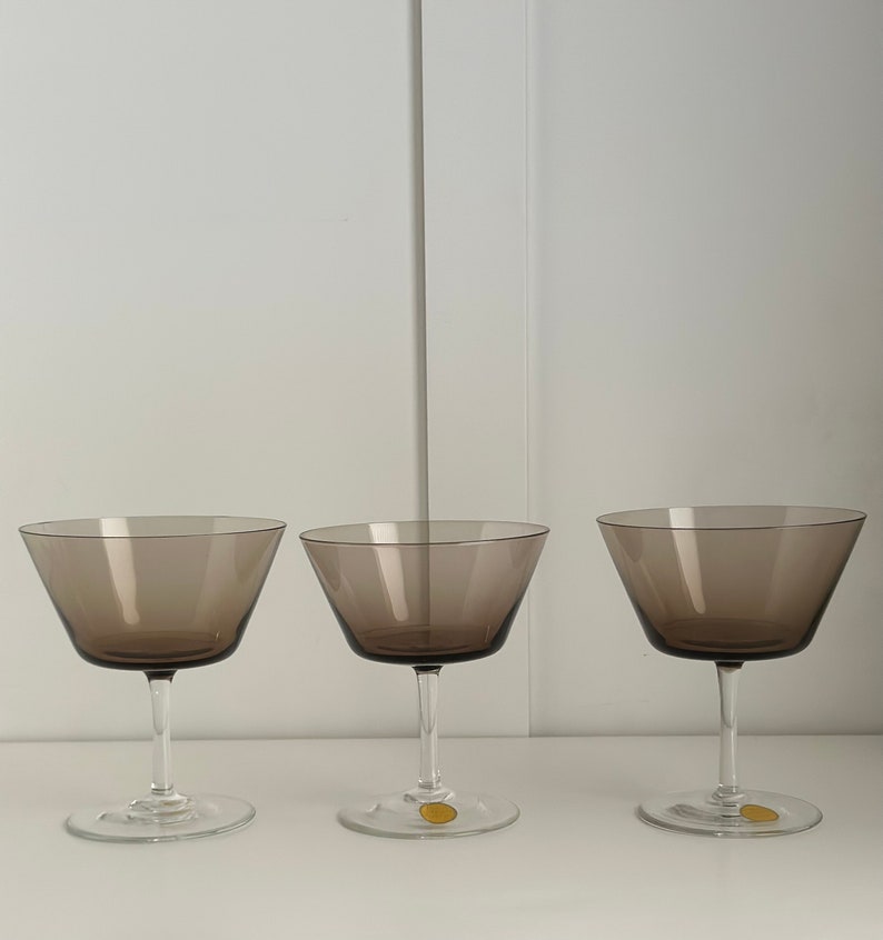 May include: Three clear glass champagne coupes with a dark brown tint. The glasses are empty and sitting on a white surface.
