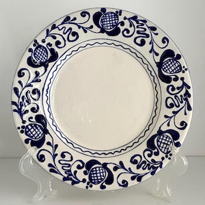 Op de afbeelding: Een witte keramische plaat met een blauw bloemmotief. De plaat heeft een herhalend ontwerp van gestileerde bloemen en wijnranken langs de rand. Het midden is effen wit en de rand heeft een geschulpte rand.