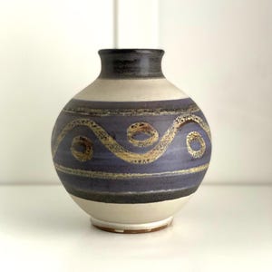 Op de afbeelding: Een keramische vaas met een afgeronde body en een smalle hals. De vaas heeft een crèmekleurige basis, een donkerblauw middengedeelte met een gele wervelontwerp en een donkergrijze rand. De vaas is een decoratief woondecoratie-item.