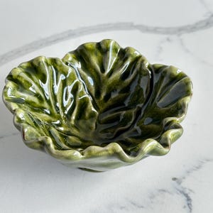 May include: A ceramic bowl shaped like a ruffled cabbage leaf with a glossy finish. The bowl features deep forest green and lighter sage green tones, with realistic molded veins and a wavy, scalloped edge that mimics the texture of a vegetable.