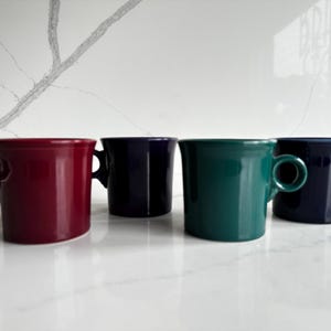Puede incluir: Un juego de cuatro tazas de cerámica en colores sólidos, incluyendo rojo intenso, morado oscuro, verde azulado vibrante y azul marino. Cada taza presenta un acabado liso y brillante, un cuerpo cilíndrico y un asa redondeada en el lateral.