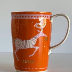 May include: Orange ceramic mug with a white handle and base. The mug features a white and pink Aries zodiac ram design and the word "ARIES" in gold lettering. The rim has a pink dotted pattern.