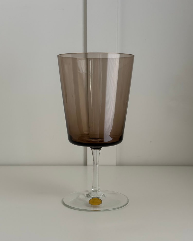 May include: A dark brown glass wine glass with a clear stem and base. The base has a gold sticker with the text "Cristal d'Arques" on it.
