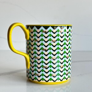 Op de afbeelding: Een keramische mok met een gele handgreep en rand. De mok heeft een herhalend zigzagpatroon in groen, zwart en lichtblauw, met gouden accenten. Een decoratief item voor dranken.