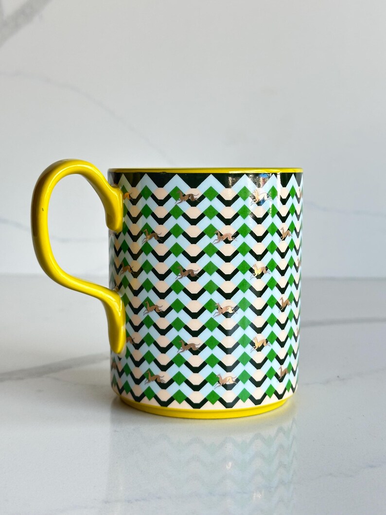 Op de afbeelding: Een keramische mok met een gele handgreep en rand. De mok heeft een herhalend zigzagpatroon in groen, zwart en lichtblauw, met gouden accenten. Een decoratief item voor dranken.