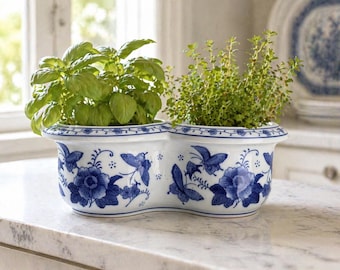 Puede incluir: Una jardinera de cerámica blanca y azul con dos secciones, cada una con una hierba diferente. La jardinera está decorada con diseños florales y mariposas azules. Las hierbas son verdes y exuberantes, añadiendo un toque natural a la decoración.