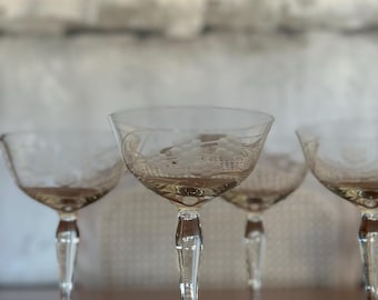May include: Four clear glass stemmed cocktail glasses with etched floral designs. The glasses are arranged in a row on a wooden surface.