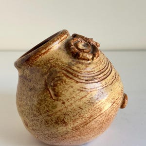 May include: A small, handcrafted ceramic vessel with a rounded body and a wide, angled opening. The pottery is a mottled tan color with darker brown accents and a textured surface. Decorative spiral and swirl patterns are visible.