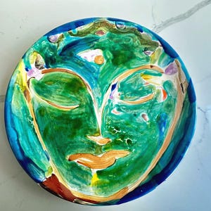 May include: A round, hand-painted ceramic plate featuring a face design. The plate has a vibrant color palette, with shades of green, blue, orange, and yellow. The face has closed eyes and a serene expression. The plate's rim is a deep blue.