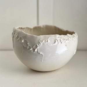 Puede incluir: Un cuenco de cerámica blanca con un borde texturizado e irregular. El exterior del cuenco presenta un delicado patrón en relieve alrededor de la parte superior. La superficie lisa y brillante contrasta con el borde texturizado.