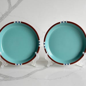 May include: Two round ceramic plates featuring a solid turquoise center and a dark brown rim. The rim is accented with small, evenly spaced blue and white rectangular patterns. Each plate is displayed upright on a clear plastic stand.