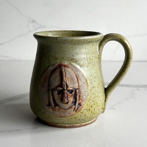 Może przedstawiać: Ceramiczny kubek z nakrapianą jasnozieloną glazurą i zakrzywionym uchwytem. Z przodu znajduje się okrągły medalion z wypukłym, ręcznie rzeźbionym reliefem twarzy w hełmie w odcieniu jasnobrązowej gliny, ukazujący szczegółowe rysy twarzy.