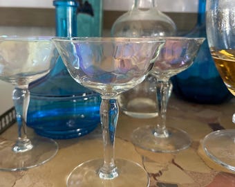 May include: Four iridescent clear glass champagne coupes on a marble surface. The coupes are arranged in a semi-circle with the front coupe in focus.