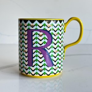 Op de afbeelding: Een keramische mok met een gele handgreep en rand. De mok heeft een paarse letter "R" op een achtergrond van groene, zwarte en witte zigzagpatronen. Kleine bruine vogelillustraties zijn over het ontwerp verspreid.