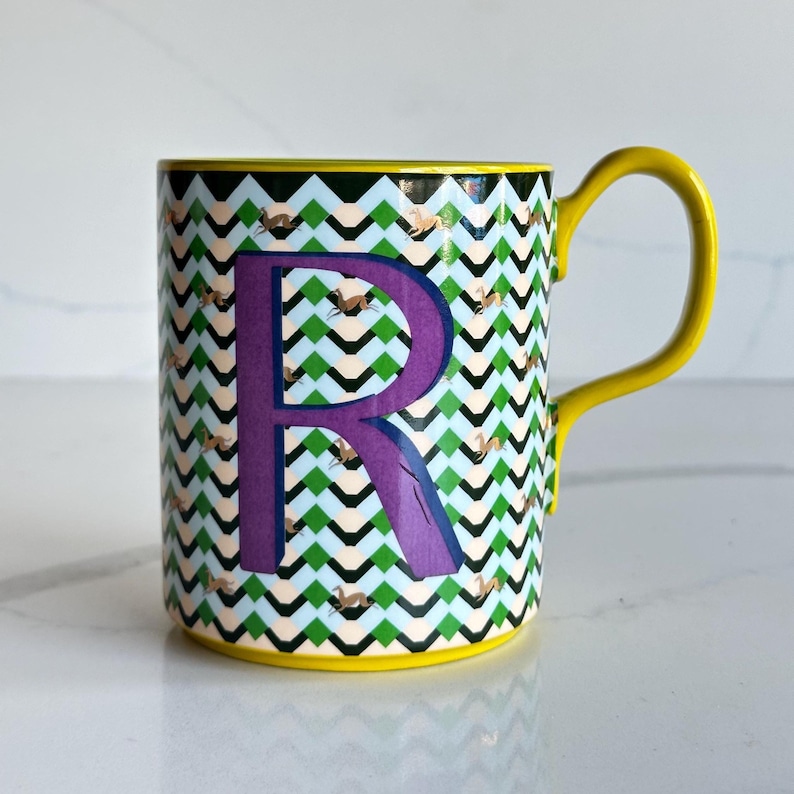 Op de afbeelding: Een keramische mok met een gele handgreep en rand. De mok heeft een paarse letter "R" op een achtergrond van groene, zwarte en witte zigzagpatronen. Kleine bruine vogelillustraties zijn over het ontwerp verspreid.