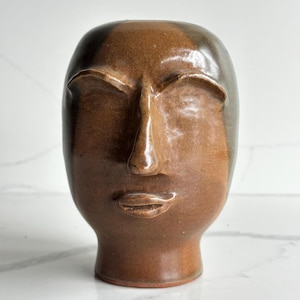 May include: A ceramic sculpture shaped like a human head with a smooth, glossy brown glaze. The face features stylized, raised eyebrows, a prominent nose, and defined lips. The surface has a reflective finish with darker brown shading.