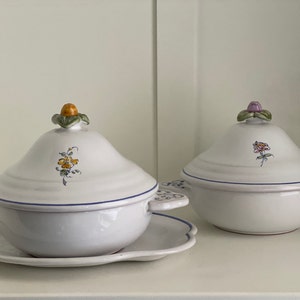 May include: Two white ceramic serving dishes with lids. The lids have floral designs and are topped with small, decorative knobs. The dishes are on a white surface.