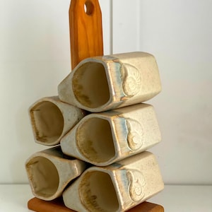Puede incluir: Un juego de cuatro servilleteros de cerámica color hueso con acabado moteado y un motivo de concha en relieve, apilados en un soporte de madera vertical. Cada anillo presenta un sutil acento de esmalte azul y marrón en los bordes.