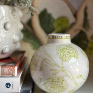 Könnte beinhalten: Eine weiße Keramikvase mit grünem Blumendesign. Die Vase ist rund und hat eine breite Öffnung. Sie steht auf einem Stapel Bücher.