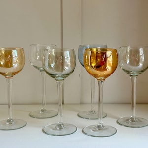 May include: Six stemmed wine glasses with round bowls. The glasses have clear stems and bases. Two glasses have a golden iridescent finish, one has a blue iridescent finish, and the other three are clear.