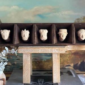 May include: A wooden box with six individual compartments, each containing a white ceramic bust of a person. The box is sitting on a wooden table.