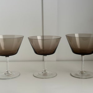 May include: Three clear glass champagne coupes with a brown tint. The coupes are arranged in a row on a white surface.