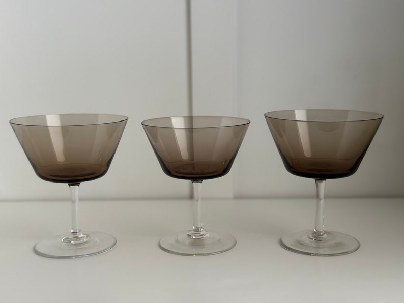 May include: Three clear glass champagne coupes with a brown tint. The coupes are arranged in a row on a white surface.