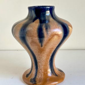 Peut inclure: Petit vase artisanal en céramique de forme unique. Il présente une base et un corps bruns avec des rayures verticales bleu marine. Le bord du vase est bleu marine. Le vase a une finition brillante et repose sur une petite base arrondie.