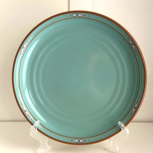 Puede incluir: Un plato redondo de cerámica con un centro de color turquesa claro sólido y un acabado liso y brillante. El borde está definido por una fina línea marrón oscuro acentuada con pequeños patrones geométricos simétricos en blanco, negro y turquesa.