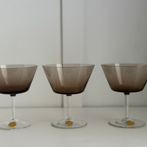 May include: Three clear glass champagne coupes with a dark brown tint. The glasses are empty and have a classic design.