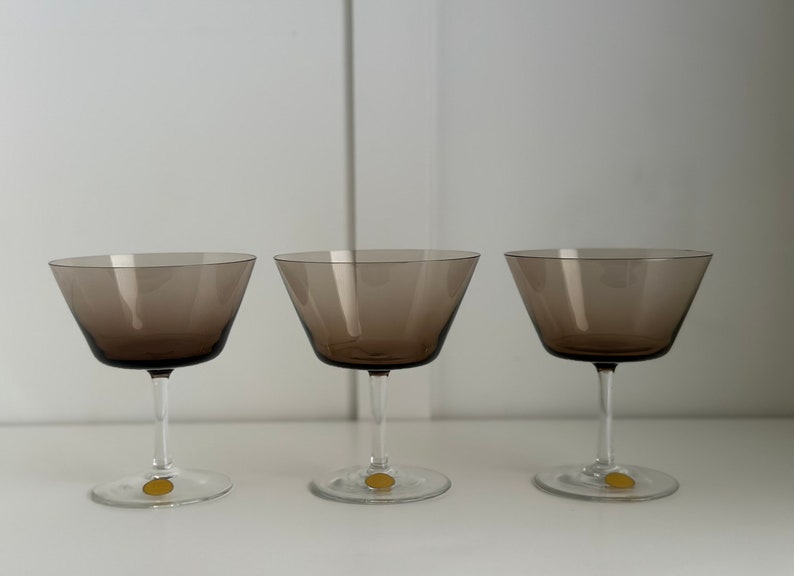 May include: Three clear glass champagne coupes with a dark brown tint. The glasses are empty and have a classic design.