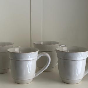 May include: Four white ceramic mugs with a subtle brown rim. The mugs have a simple design with a single band around the middle.