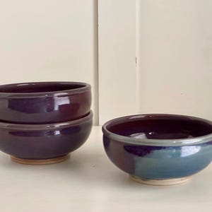 May include: Three ceramic bowls in shades of purple and blue. Two bowls are stacked, and the third is separate. The bowls have a glossy finish and sit on a light-colored base. The bowls are suitable for serving food.