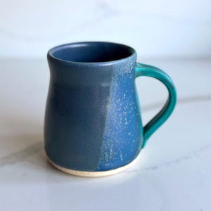 Puede incluir: Taza de cerámica hecha a mano con un esmalte azul de dos tonos. La taza presenta un cuerpo azul oscuro que transiciona a un azul más claro con detalles moteados. El asa es de color turquesa contrastante y la base no está esmaltada. Ideal para café o té.