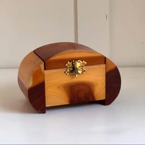May include: A wooden jewelry box with a curved top and a rectangular front. The box is made of light and dark brown wood, with a brass clasp. The box is on a white surface.