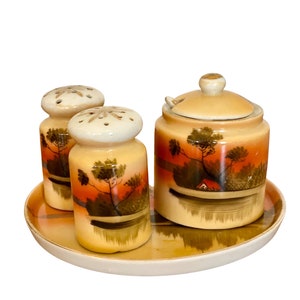 Może przedstawiać: Zestaw vintage solniczki i pieprzniczki ceramicznych z pasującą śmietanką i tacą. Zestaw zawiera ręcznie malowany krajobraz z drzewami, domem i jeziorem. Elementy są w odcieniach brązu, pomarańczy i żółci.