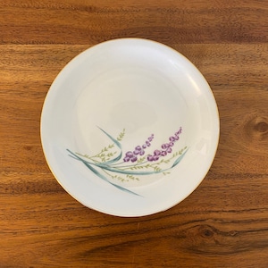 May include: A white ceramic plate with a gold rim features a delicate floral design. The design includes purple flowers and green leaves.