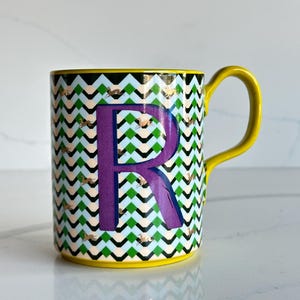 Op de afbeelding: Een keramische mok met een gele handgreep en rand. De mok heeft een zigzagpatroon in groen, zwart en wit, met gouden accenten. Een grote paarse letter "R" staat prominent op de voorkant.