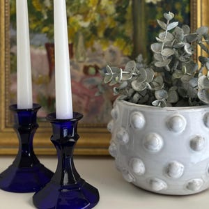 May include: Two white candles in dark blue glass candle holders. A white ceramic pot with a textured surface and a plant with gray-green leaves is to the right. A framed painting is in the background.
